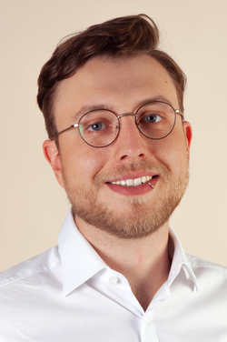 Nahaufnahme eines lächelnden jungen Mannes mit braunen Haaren, blauen Augen, Brille, Bart und Lippenpiercing, in weißem Hemd.