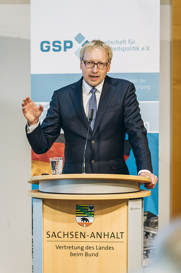 Mann mit blonden Haaren und Brille im dunklen Anzug spricht an einem Rednerpult mit Logos von 'Sachsen-Anhalt' und 'GSP'.