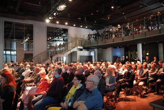 Großes Publikum von Erwachsenen in einem modernen Auditorium mit Mezzanin, das eine Veranstaltung oder Konferenz besucht.