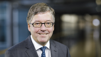 Lächelnder Mann mit grauem Haar und Brille, Anzug und blauer Krawatte, vor einem unscharfen modernen Hintergrund.