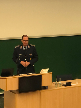 Ein uniformierter Offizier steht an einem Rednerpult in einem Hörsaal mit einer grünen Tafel.
