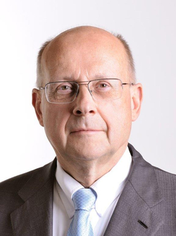 Porträt eines Glatzkopfes mit Brille, grauem Anzug und hellblauer Krawatte vor schlichtem weißem Hintergrund.