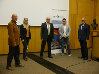 Fünf Personen, darunter Männer und eine Frau, stehen lächelnd in einer Reihe bei einer Veranstaltung mit einem GSP-Banner.