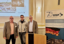 Drei Männer bei einer Veranstaltung der Gesellschaft für Sicherheitspolitik (GSP) mit Katastrophenbildern auf dem Bildschirm und GSP-Banner.