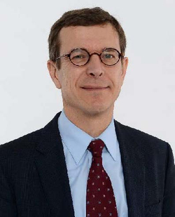 Porträt eines Mannes in dunklem Anzug, hellblauem Hemd und roter Krawatte, mit runder Brille lächelnd vor weißem Hintergrund.