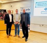 Drei lächelnde Männer bei einem Event mit Präsentationsbildschirm, der Text und ein '70 BSP'-Banner zeigt.