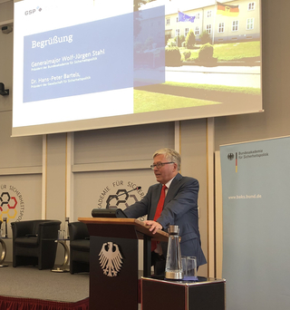Mann am Rednerpult mit Bundesadler. Leinwand zeigt 'Begrüßung' für Veranstaltung der Bundesakademie für Sicherheitspolitik.
