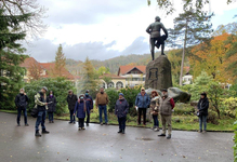 Eine Gruppe mit einem Führer in einem Herbstpark, stehend nahe einer Statue auf einem Felsen, Brunnen und Gebäude im Hintergrund.