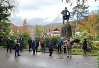 Eine Gruppe mit einem Führer in einem Herbstpark, stehend nahe einer Statue auf einem Felsen, Brunnen und Gebäude im Hintergrund.