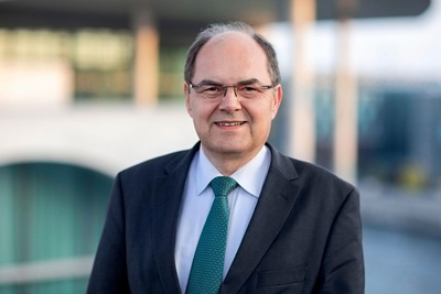 Lächelnder Mann mit Brille in dunklem Anzug und grünem Krawatte, draußen vor verschwommenem modernen Gebäudehintergrund.