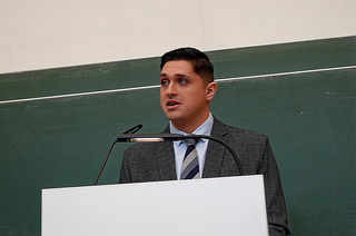 Ein junger Mann im Anzug spricht an einem Pult mit Mikrofon vor einer Tafel.