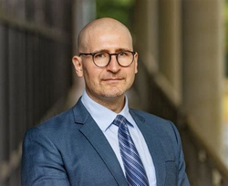 Ein glatzköpfiger Mann mit Brille und Bart, im blauen Anzug und gestreifter Krawatte, lächelt in die Kamera.