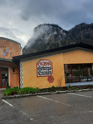 Ein oranges Gebäude mit 'Grassl Gebirgs Enzian' Schild, dahinter ein nebelverhangener Berg und ein Parkplatz.