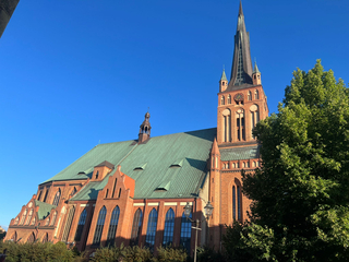 Eine große gotische Backsteinkirche mit hohem, dunklem Turm und grünem Dach vor blauem Himmel, flankiert von grünen Bäumen.