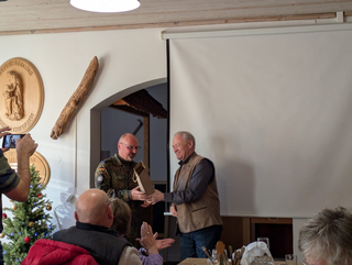 Ein lächelnder Mann in Militäruniform überreicht einem lächelnden Mann in Weste eine Geschenktüte bei einer Veranstaltung mit Zuschauern.
