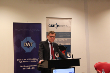 Ein Mann im Anzug spricht an einem Rednerpult vor Bannern der DWT (Wehrtechnik) und GSP (Sicherheitspolitik).