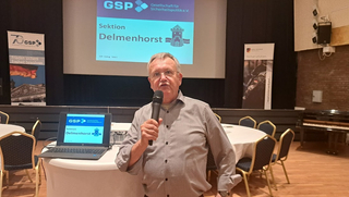 Mann spricht mit Mikrofon bei GSP-Veranstaltung, mit „Sektion Delmenhorst“ auf Leinwand und Bannern.