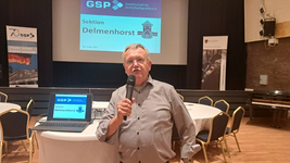 Mann spricht mit Mikrofon bei GSP-Veranstaltung, mit „Sektion Delmenhorst“ auf Leinwand und Bannern.