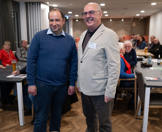 Zwei lächelnde Männer bei einer Veranstaltung, einer mit Namensschild, umgeben von sitzenden Gästen.