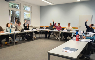 Junge Erwachsene in einem Klassenzimmer, einige heben an einem U-förmigen Tisch die Hände, was auf eine Abstimmung oder Diskussion hindeutet.