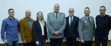 Gruppenfoto von sieben Personen: zwei Männer in Militäruniformen und fünf Zivilisten (drei Männer, eine Frau) stehen lächelnd in einer Reihe.