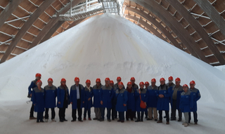 Gruppe in blauen Mänteln und Helmen in einer riesigen Industriehalle mit großem weißem Schüttguthaufen und Holzbogen-Decke.