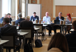 Konferenz in einem Raum mit Teilnehmern an Tischen, einige mit Laptops. Eine rothaarige Frau lächelt.