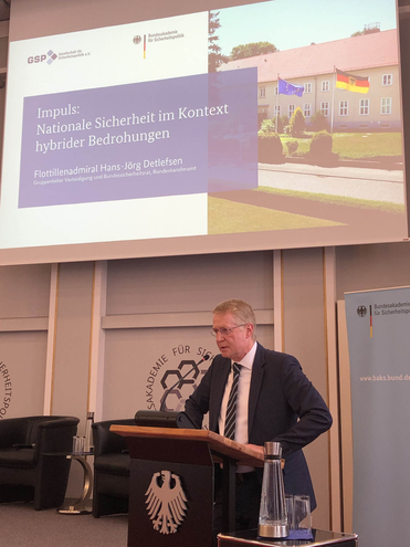 Mann spricht am Rednerpult. Präsentation: 'Nationale Sicherheit im Kontext hybrider Bedrohungen' von Flottillenadmiral Detlefsen.
