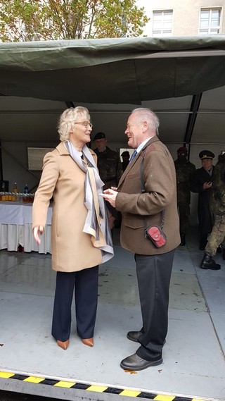 Ursula von der Leyen, im beigen Mantel, lächelt herzlich im Gespräch mit einem Mann in brauner Jacke bei einer Veranstaltung.