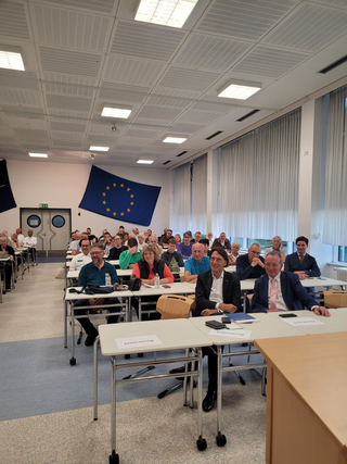 Teilnehmer eines Meetings in einem Raum mit EU-Flaggen und Reihen von Tischen, einige Personen lächeln.