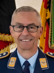 Ein lächelnder deutscher Militäroffizier mit Brille und grauem Haar in dunkelblauer Uniform. Deutsche Flaggen im Hintergrund.