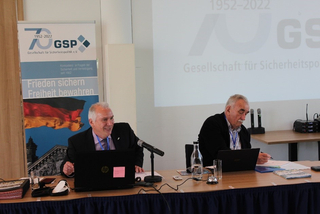 Zwei Männer bei einer GSP-Konferenz. Einer lächelt, der andere schreibt. Ein Banner der Gesellschaft für Sicherheitspolitik ist dahinter.