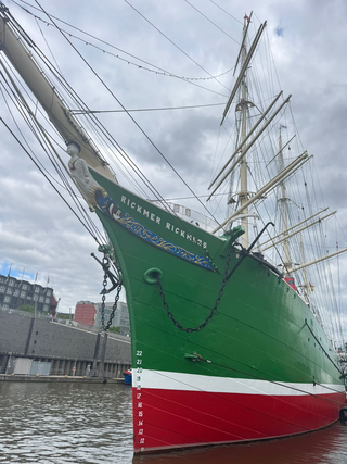 Das grüne Segelschiff Rickmer Rickmers mit rotem Rumpf und Galionsfigur am Bug liegt im Hafen unter bewölktem Himmel.