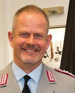 Lächelnder Mann mit kurzen grauen Haaren und Spitzbart in hellgrauer Militäruniform mit roten Schulterabzeichen.