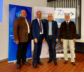 Vier Männer, teils in Anzügen, teils in Trachtenjacken, lächeln vor Roll-up-Bannern deutscher Organisationen.