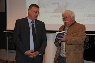 Zwei Männer bei einer Buchpräsentation: Einer im Anzug, der andere in Trachtenjacke hält ein Buch vor einer Projektionsfläche.