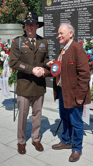 Lächelnder Soldat in Kavalleriehut schüttelt älterem Zivilisten mit Plakette an Militärdenkmal die Hand.