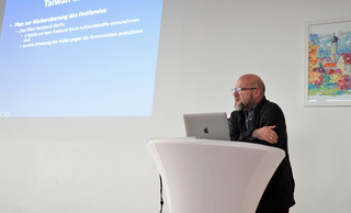 Mann präsentiert mit Laptop vor einer Leinwand mit Text über "Plan zur Rückeroberung des Festlandes", daneben ein farbiges Bild.