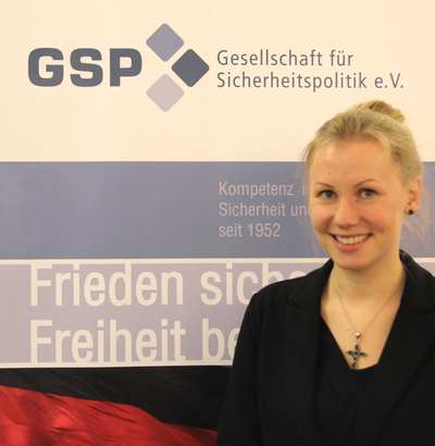 Lächelnde blonde Frau vor GSP-Banner mit Text 'Gesellschaft für Sicherheitspolitik e.V.', 'Kompetenz in Sicherheit seit 1952' und deutscher Flagge.