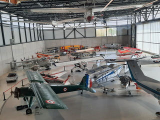 Museumshangar mit vielen historischen Flugzeugen und Segelflugzeugen, einige hängen, andere am Boden, und einem Oldtimer.