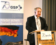 Mann spricht auf GSP-Veranstaltung (70. Jubiläum) mit Banner 'Frieden sichern Freiheit bewahren' und deutscher Flagge.