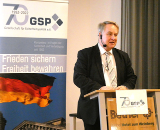 Mann spricht auf GSP-Veranstaltung (70. Jubiläum) mit Banner 'Frieden sichern Freiheit bewahren' und deutscher Flagge.
