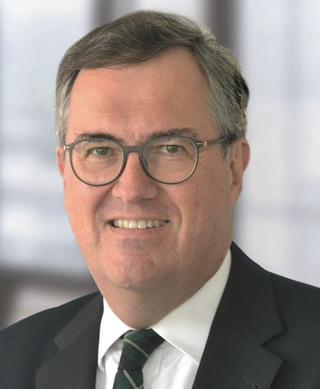 Porträt eines lächelnden Mannes mit grauem Haar und Brille, der einen dunklen Anzug und eine grün gestreifte Krawatte trägt.