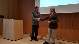 Ein Mann in Militäruniform und ein Mann im Blazer schütteln sich die Hände. Im Hintergrund ein Rednerpult und eine Projektionsfläche.