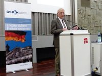 Älterer Mann spricht an Rednerpult mit GSP-Banner, das deutsche Flagge und Reichstagsgebäude zeigt.