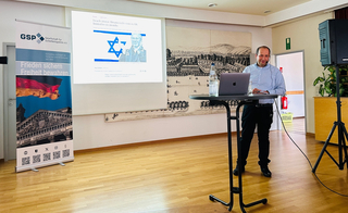 Ein Mann präsentiert mit Laptop, eine Leinwand zeigt israelische Flaggennachrichten, daneben ein GSP-Banner.