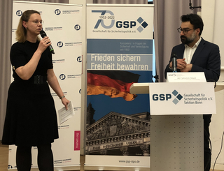 Eine Frau spricht in ein Mikrofon neben einem Mann an einem Podium, mit Bannern der GSP und Friedrich Naumann Stiftung.