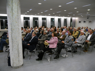 Ein Publikum aus hauptsächlich Erwachsenen, Männern und Frauen, die in Reihen in einer Innenhalle mit einer dekorativen Fliesensäule sitzen.