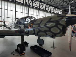 Ein getarntes Messerschmitt Bf 108 Taifun Propellerflugzeug mit Emblem am Rumpf in einem hellen Hangar.