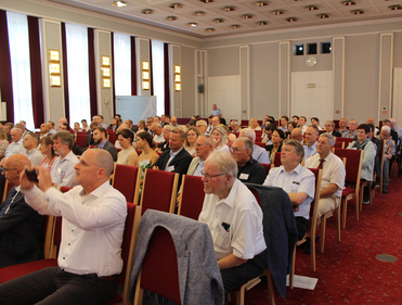 Ein vielfältiges erwachsenes Publikum sitzt in Reihen roter Stühle in einem Konferenzsaal, ein Mann fotografiert.